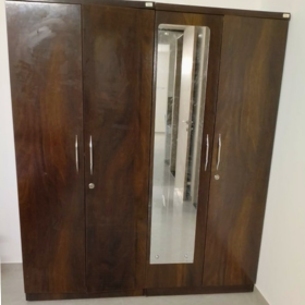 A dark brown wooden four-door large wardrobe storage option with two doors featuring vertical handles and one central door with a large rectangular mirror, set against a plain white wall. Available on affordable monthly rent with free delivery in pune kharadi wagholi Hadapsar Amanora Magarpatta keshav nagar Viman Nagar Aundh Baner Balewadi Hinjewadi Wakad ravel punawale.