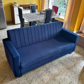 A modern three-seater sofa in deep navy blue fabric, featuring vertical channel tufting on the backrest, positioned in a dining room with a dining table and chairs in the background. Available on affordable monthly rent with free delivery in pune kharadi wagholi Hadapsar Amanora Magarpatta keshav nagar Viman Nagar Aundh Baner Balewadi Hinjewadi Wakad ravel punawale.