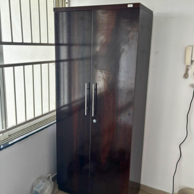 A tall dark brown wooden two-door cabinet with silver handles, standing in a room next to a barred window. delivered in pune kharadi wagholi Hadapsar Amanora Magarpatta keshav nagar Viman Nagar Aundh Baner Balewadi Hinjewadi Wakad ravel punawale.